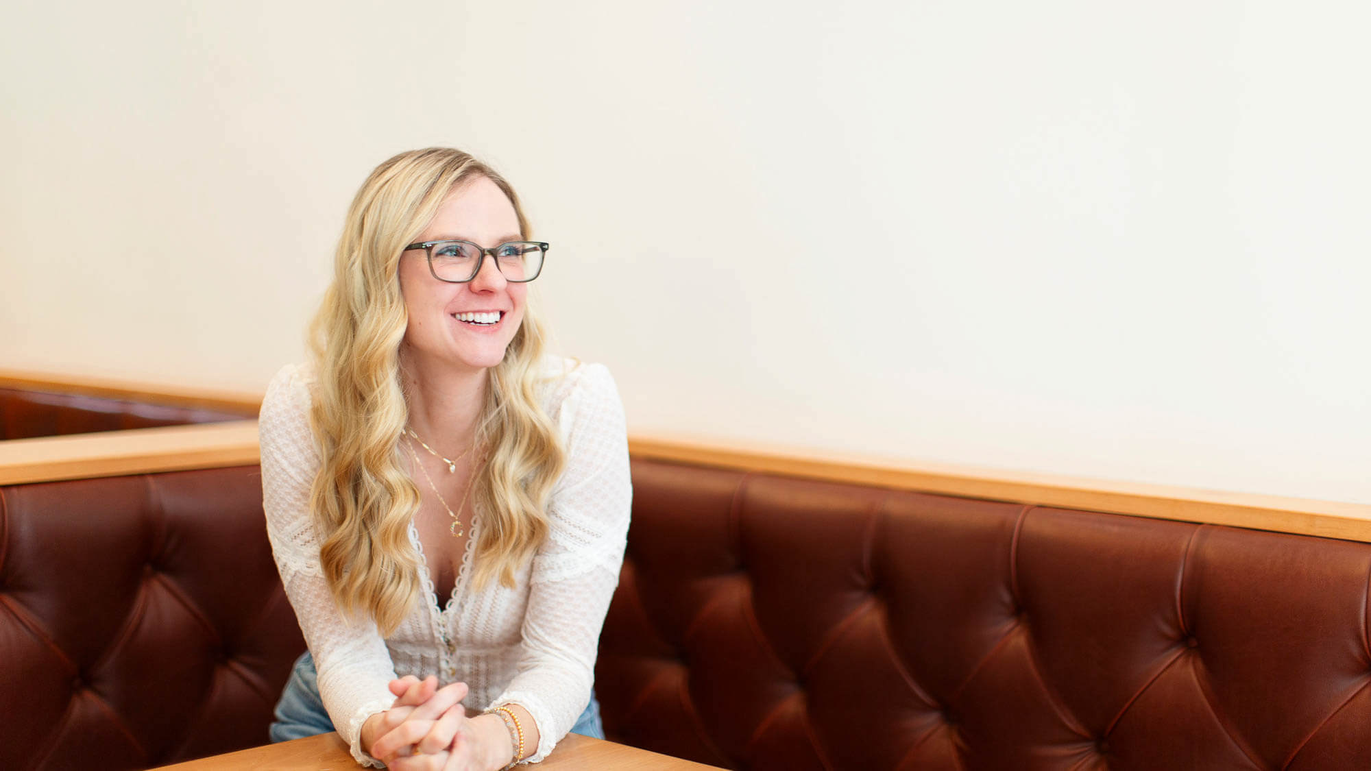 Smiling blonde jeweler in glasses seated in a booth, white blouse and layered necklaces — Sara Gallo Custom Jewelers, Lancaster, PA.